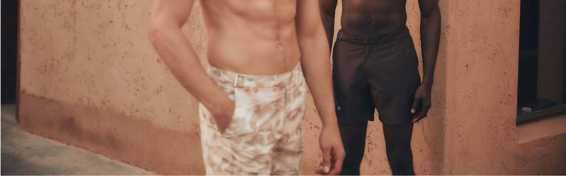 Two men standing against a beige wall, wearing luxury swim shorts brand called lewis james